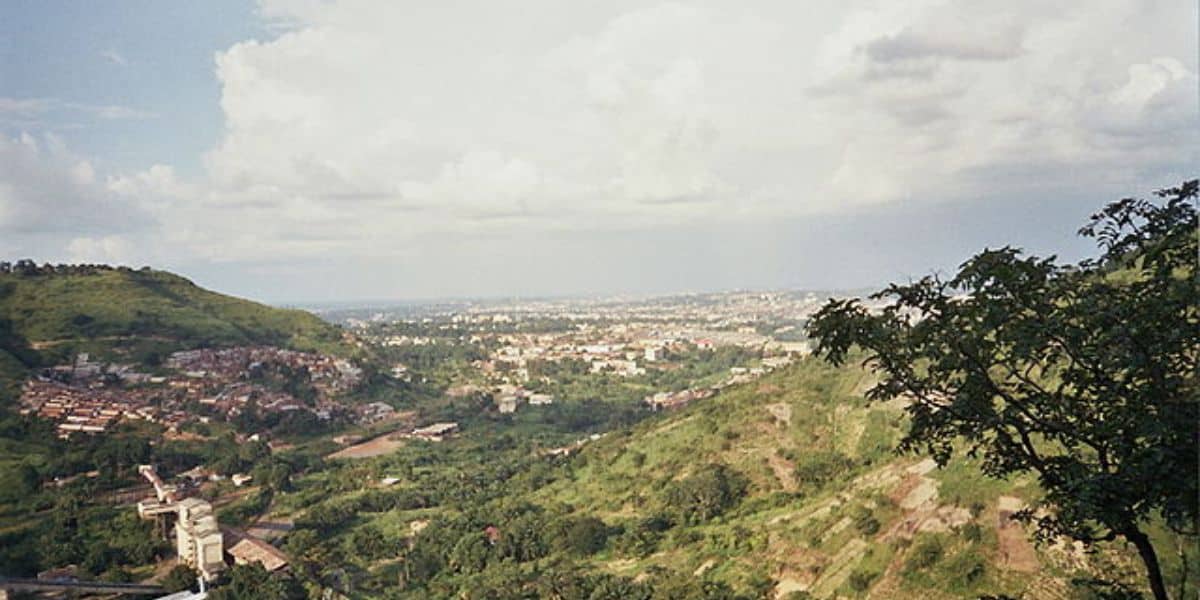 A panoramic view of a city surrounded by lush greenery and hills under a partly cloudy sky. The cityscape extends into the distance, blending with the natural landscape of rolling hills and dense vegetation.