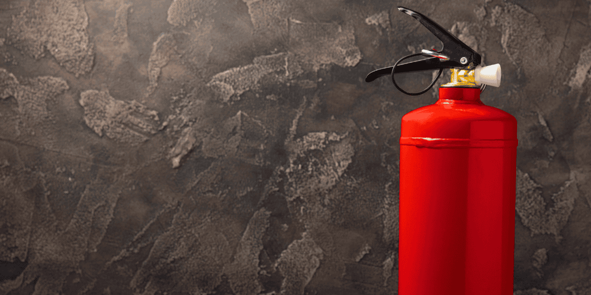 A red fire extinguisher with a black nozzle and safety pin is positioned against a textured, dark brown wall. The stucco-like finish, reminiscent of Taylor Swift's home decor style, adds a rugged backdrop to the simple, bright extinguisher.