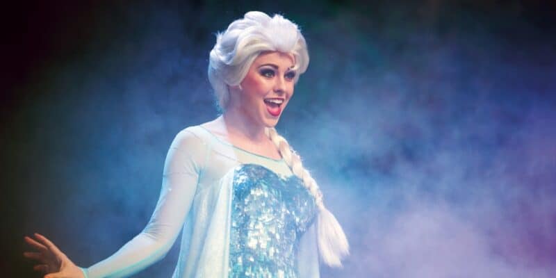 A performer dressed as an ice queen with light blonde hair in a braid, wearing a sparkling blue and white gown, stands in front of a misty background. She has an enthusiastic expression and holds one hand out to the side.