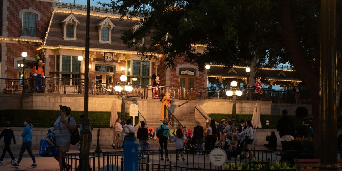 A lively evening scene at a Disney theme park entrance, featuring beautifully lit buildings adorned with lights. Characters in costumes interact with guests, some of whom are wearing masks. The area is filled with people strolling and taking in the festive atmosphere.