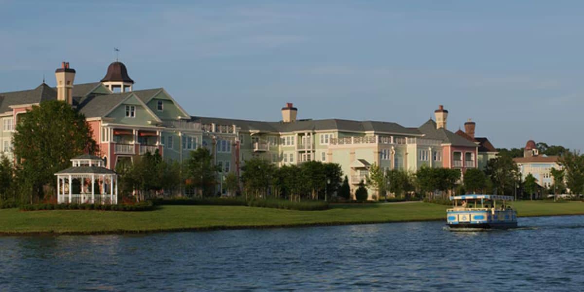 A serene lake scene with a charming, multi-colored building complex in the background, featuring pastel hues reminiscent of Disney Resort pools. A gazebo is on the left near the water's edge. A blue and yellow boat floats on the calm lake to the right. Trees and greenery surround the area.