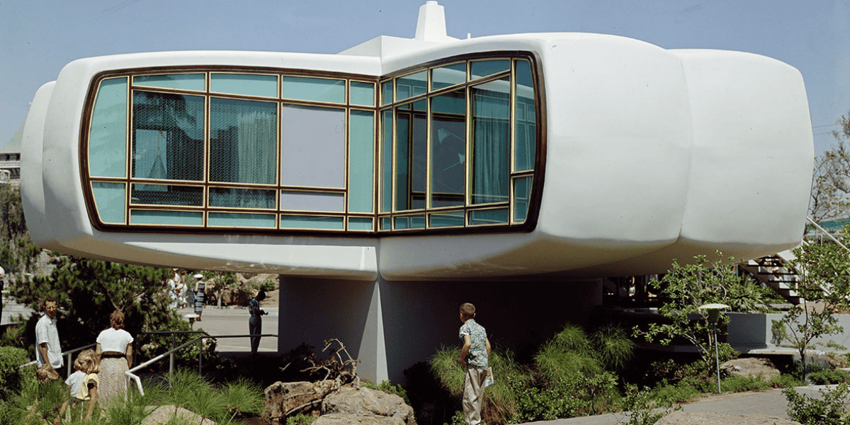 A futuristic house with a smooth, white exterior and large, rectangular windows, elevated on a central support. A family with children stands at the base, surrounded by greenery and rock landscaping. With its retro, space-age design, the home evokes a sense of Disneyland's imaginative charm.