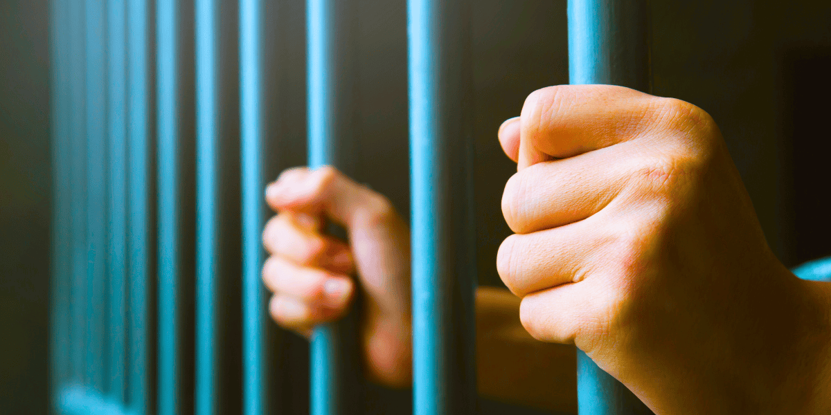 Close-up of two hands gripping the bars of a prison cell. The image highlights the stark and strong metal bars, suggesting confinement. The lighting casts a somber, reflective mood over the scene—an evocative portrayal often seen in stories of those convicted for crimes like illegal streaming on Jetflicks.