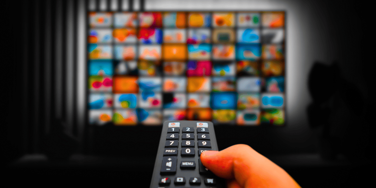 A hand holding a remote control points towards a colorful, blurry TV screen filled with numerous thumbnail images, suggesting a selection of streaming content. In the background, the dark setting underscores the vibrant screen. Recently, five men were convicted for operating Jetflicks, an illegal streaming service.