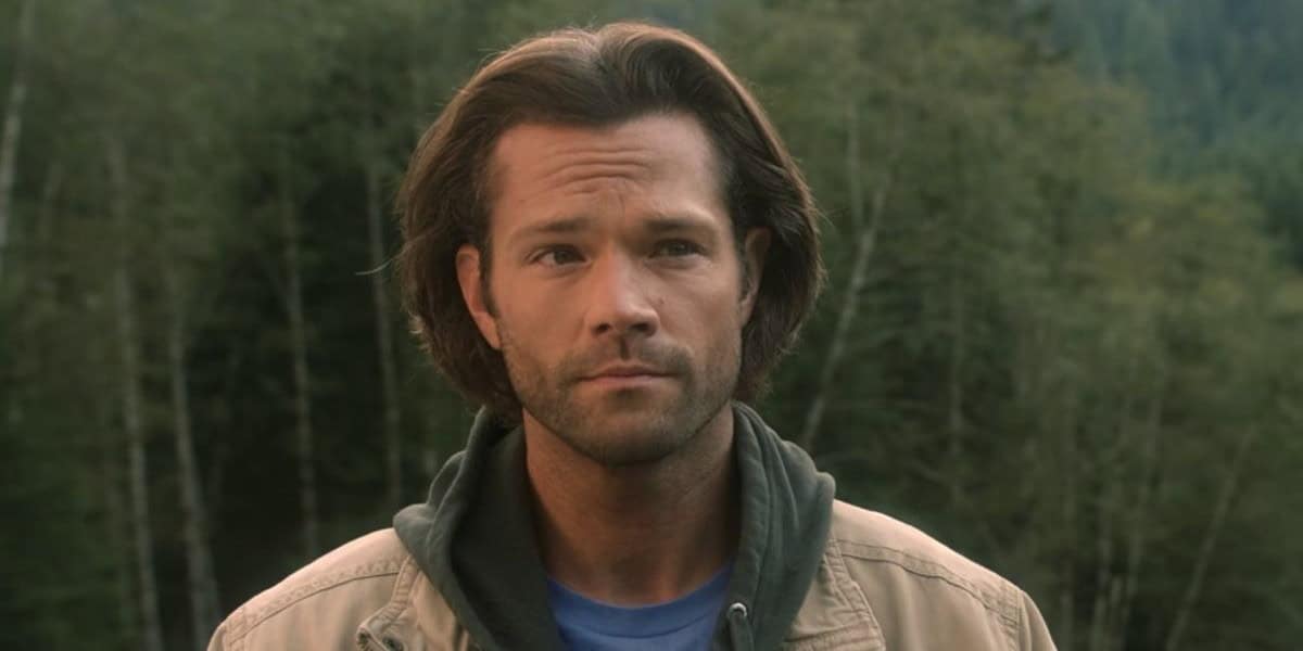 Jared Padalecki with medium-length, brown hair and facial hair is standing outdoors in front of a forested area. They are wearing a light brown jacket over a green hoodie and a blue shirt. Their expression is neutral, and they are looking slightly to the side.