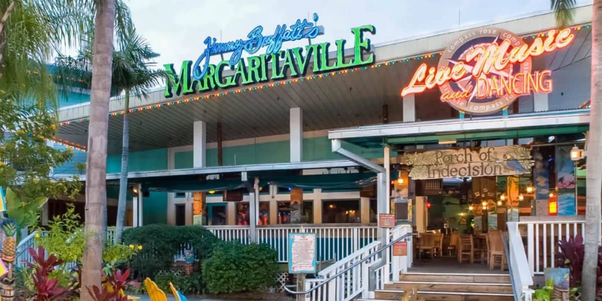A colorful outdoor view of Jimmy Buffett's Margaritaville at Universal Orlando. The building features a mix of teal and white exterior, a staircase leading to the entrance, vibrant plants, and two neon signs reading "Live Music and Dancing" and "Porch of Indecision.