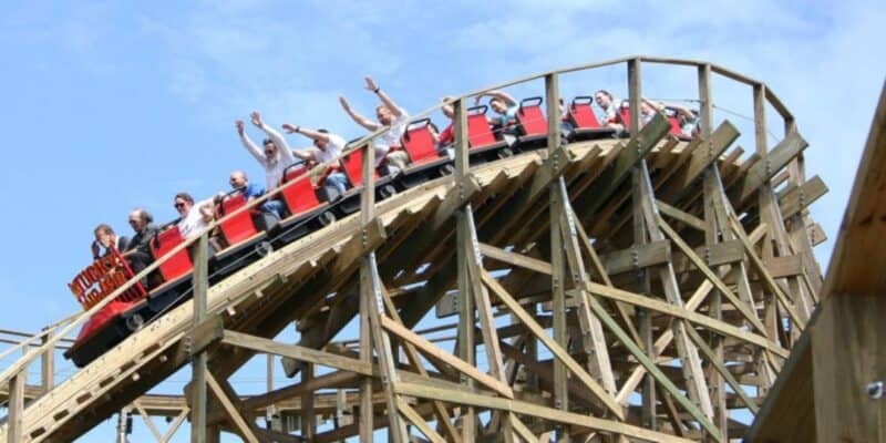 A red roller coaster filled with excited riders raises their hands as they descend a steep wooden track on a sunny day. The wooden support structure of the roller coaster is prominently visible against a clear blue sky.