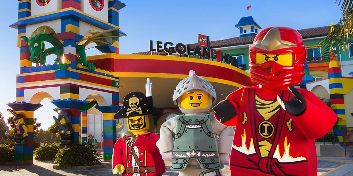 Three life-sized LEGO character figures, including a red ninja, a knight, and a pirate, stand in front of the colorful entrance to LEGOLAND Theme Park. The background features a large dragon statue on top of a multicolored building with the LEGOLAND sign.