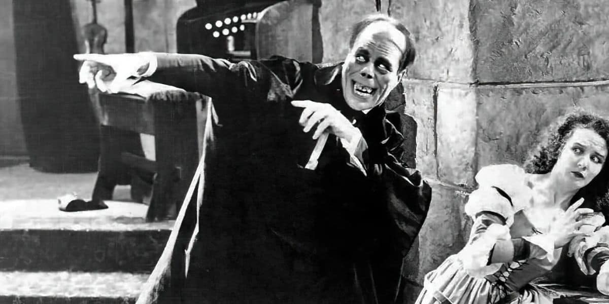 A black-and-white scene from a classic film depicts a bald man with a dramatic expression, wearing a cape and pointing forward, reminiscent of the Phantom of the Opera. A startled woman in period costume crouches nearby, looking in the direction he is pointing.