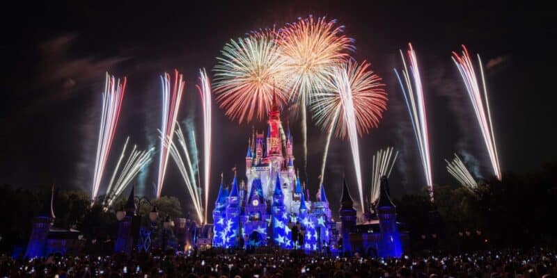 A dazzling fireworks display illuminates the night sky above a large, brightly lit castle. The castle is adorned with colorful lights and star shapes, surrounded by a crowd of spectators watching the vibrant spectacle at Magic Kingdom.