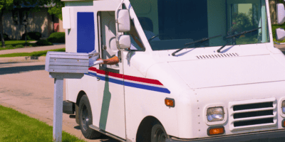 A mail carrier in a white postal truck with red, blue, and white stripes places mail into a residential mailbox on the side of a suburban street. The truck is parked on the street next to the mailbox, its door open in the bright daylight. You can almost imagine Matthew Perry driving by with a friendly wave.