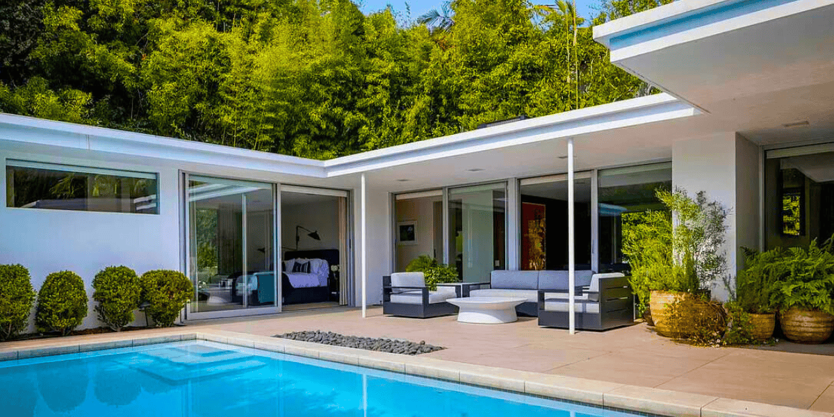 A modern house with large glass doors opens onto a poolside patio area. The patio features sleek outdoor furniture, potted plants, and a rectangular swimming pool. In the background, there is lush greenery, creating a serene and secluded atmosphere amid the recent sale of assets.