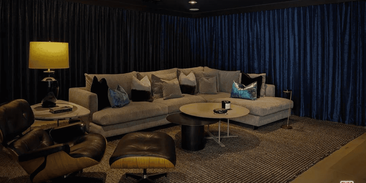 A cozy living room features a large gray sectional sofa adorned with various throw pillows. A modern lounge chair and ottoman sit nearby. Two round coffee tables are in front of the sofa, and a table lamp and decorative items complete the scene. Blue curtains, reminiscent of actor's assets, frame the backdrop.