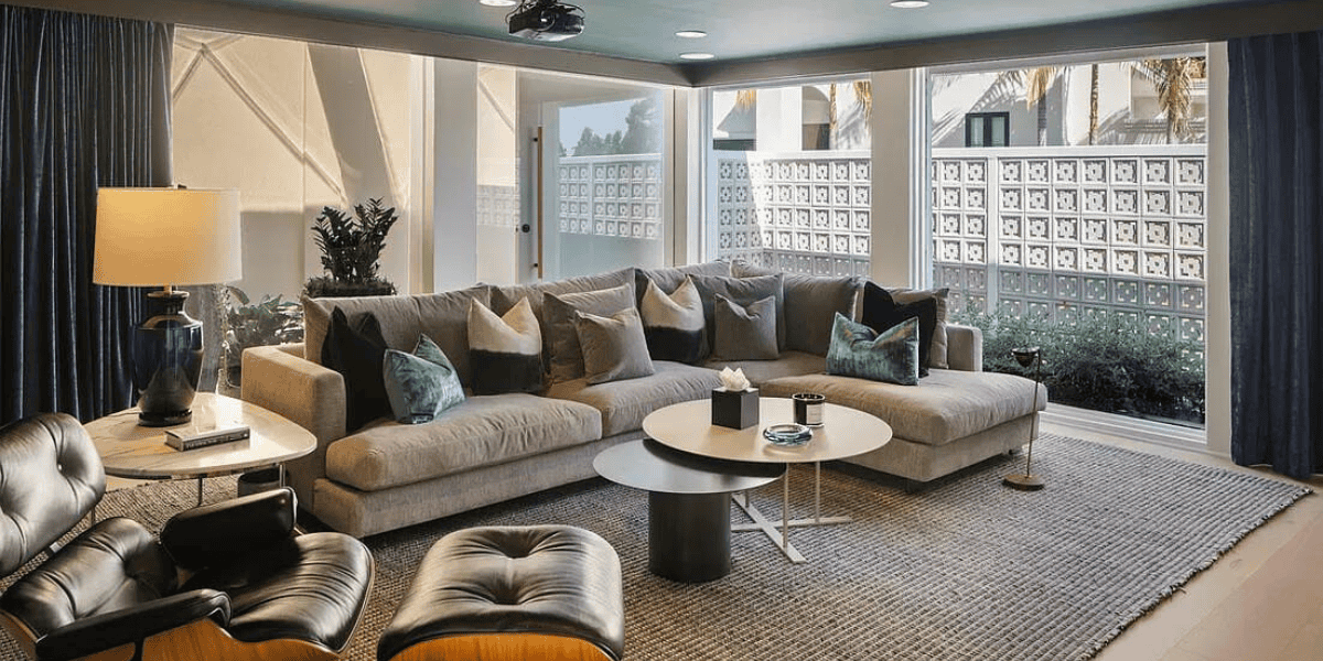 A contemporary living room features a large L-shaped grey sectional sofa with blue and green throw pillows. There's a modern round coffee table, two black leather chairs with ottomans, and a stylish lamp. Large windows provide ample natural light and garden views, setting the perfect scene for discussing any recent sale of assets.