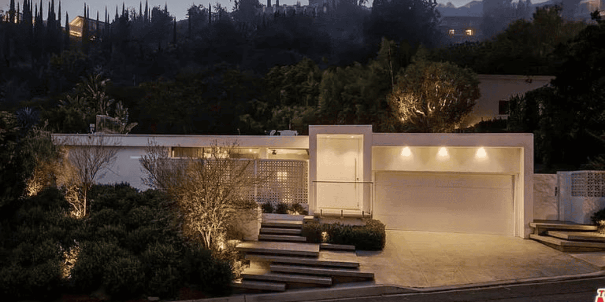 A modern, single-story house with large windows and a flat roof, surrounded by lush greenery. The exterior is illuminated with warm lighting, and steps lead up to the entrance which features a white garage door and a minimalist design. Ideal for an asset sale due to its impeccable condition.