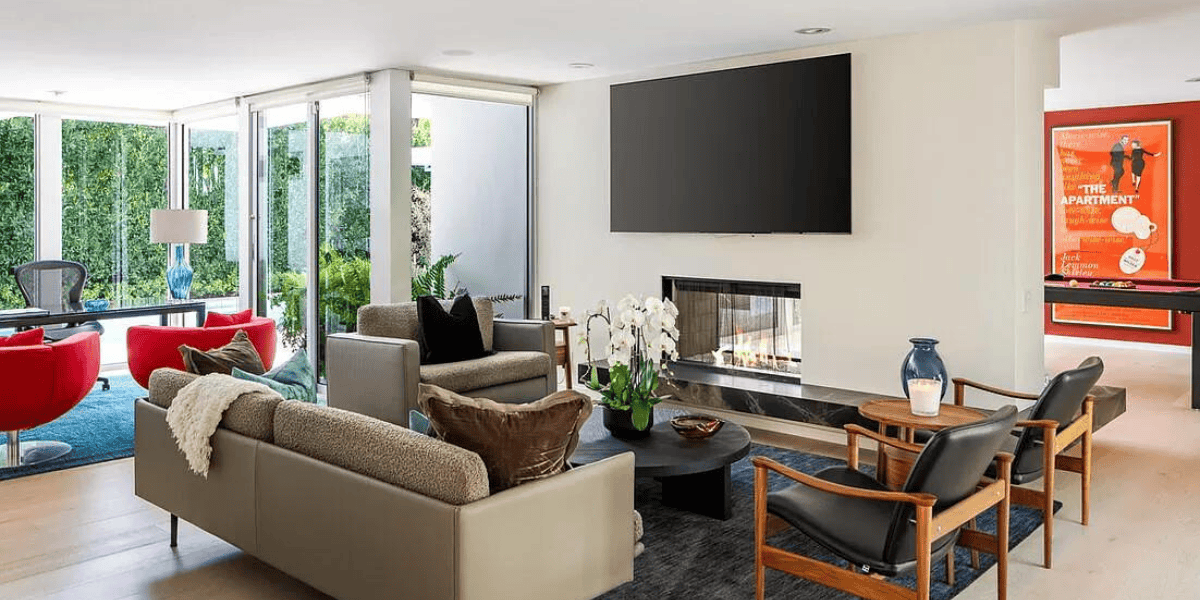 A modern, bright living room features a large, wall-mounted TV above a sleek fireplace. The room is furnished with contemporary sofas and armchairs surrounding a black coffee table with white orchids. Large windows offer garden views, and assets from Matthew Perry's estate are showcased in the background.