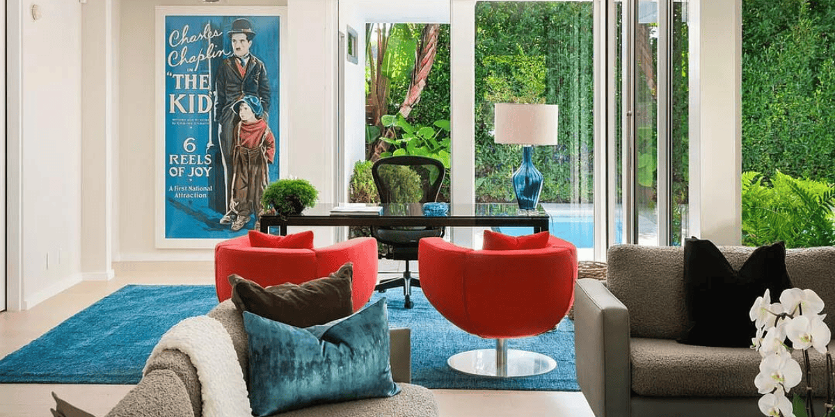 A modern living room with large windows offering a garden view. The room features a sofa, two red chairs, a desk with a chair and a lamp, and a poster of Charlie Chaplin's "The Kid" on the wall. There is a blue cushion on the sofa and a blue rug on the floor, subtly nodding to actor Matthew Perry’s distinct style.