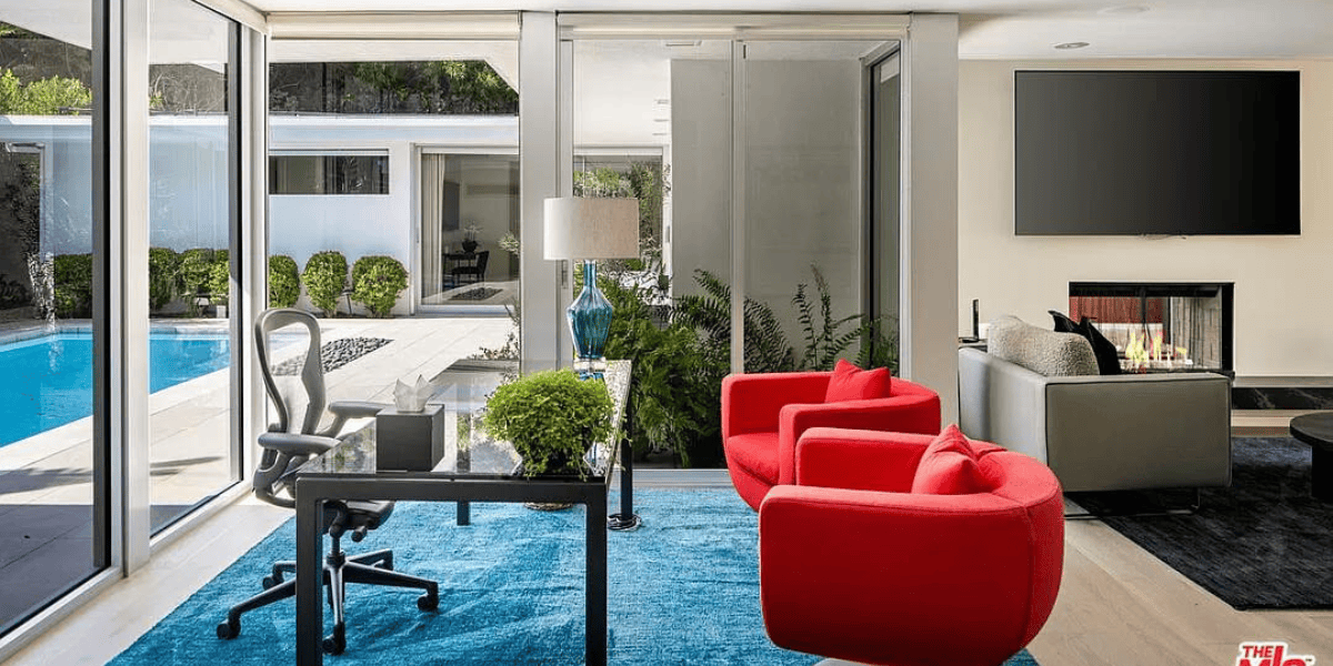 Modern living room with floor-to-ceiling glass windows overlooking a pool. Features a glass desk with a black chair, red armchairs, and a blue rug. A flat-screen TV is mounted above a fireplace. A large vase with flowers and a lamp sit on the desk. Note: house part of sale of assets.