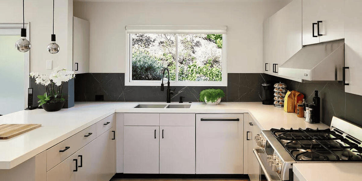 A modern kitchen with white cabinets, black countertops, and stainless steel appliances. There is a potted plant near the window, a cutting board on the counter, and an arrangement of colorful utensils next to the stove. Recently acquired in an assets sale, it basks in natural light.