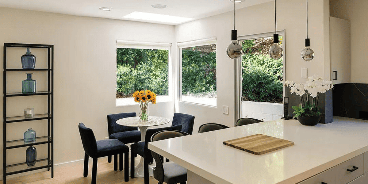 A modern kitchen and dining area with large windows overlooking lush greenery. The room features a white island countertop with a cutting board, a small round dining table with sunflowers, black chairs, hanging pendant lights, and a shelving unit adorned with blue glass bottles among an actor's assets.