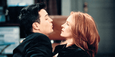 A man with dark hair, reminiscent of a young Matthew Perry, leans in for a kiss, while a red-haired woman who could be mistaken for a female celebrity leans slightly away, looking unimpressed. They are both dressed in dark clothing and seem to be in a room with bookshelves and a desk in the background.