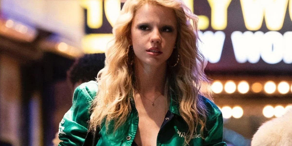 A person with long blonde hair and hoop earrings wearing a green jacket stands outdoors at night. Neon lights and signage in the blurred background create a lively atmosphere.