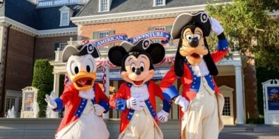 Donald Duck, Mickey Mouse, and Goofy are dressed in red, white, and blue colonial costumes, standing in front of a building with a sign that reads "The American Adventure."