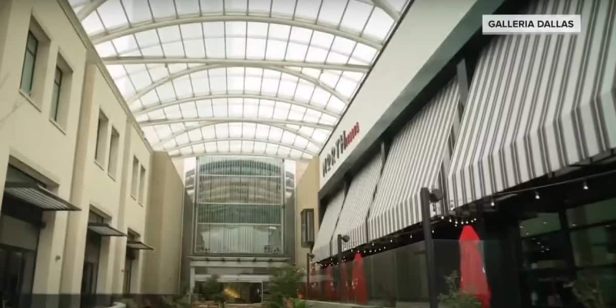 Mall interior with a glass ceiling allowing natural light, featuring multiple storefronts and a sign reading "Galleria Dallas" in the top right corner. The right side has a prominent striped awning with red umbrellas beneath. Plants line the walkway.