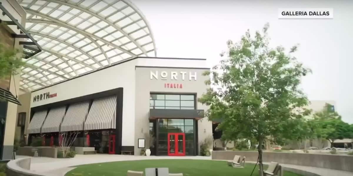 A modern building with a curved glass roof houses a restaurant named "North Italia." The entrance features large windows and bright red framed doors. Green landscaping with trees and seating surrounds the entrance. A sign indicates the location, "Galleria Dallas.