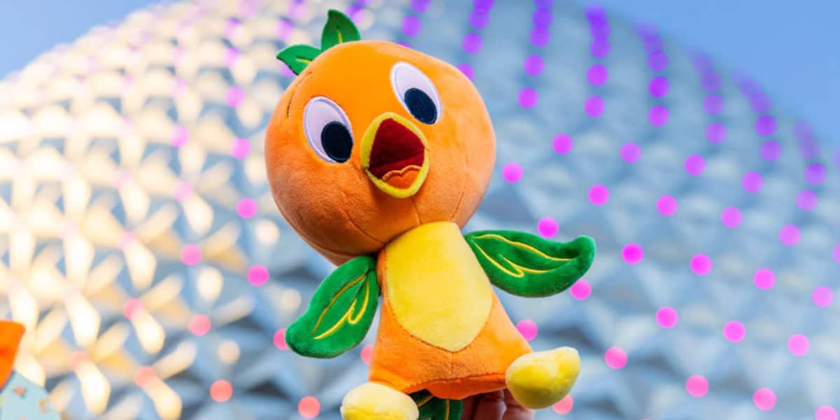 A plush toy shaped like an orange bird with green wings and leaves on its head is held up in front of a geodesic dome structure decorated with pink lights. The bird has a happy, open-mouthed expression.