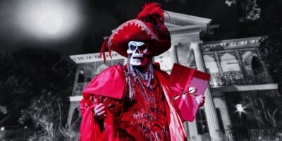A person dressed in a red skeleton costume stands in front of an old mansion at night, reminiscent of the Phantom of the Opera. The costume includes an elaborate hat with feathers, a mask with a skull design, a flowing cloak, and jewelry. The person is holding a red gift box tied with a gold ribbon.