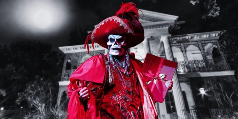 A person dressed in a red skeleton costume stands in front of an old mansion at night, reminiscent of the Phantom of the Opera. The costume includes an elaborate hat with feathers, a mask with a skull design, a flowing cloak, and jewelry. The person is holding a red gift box tied with a gold ribbon.