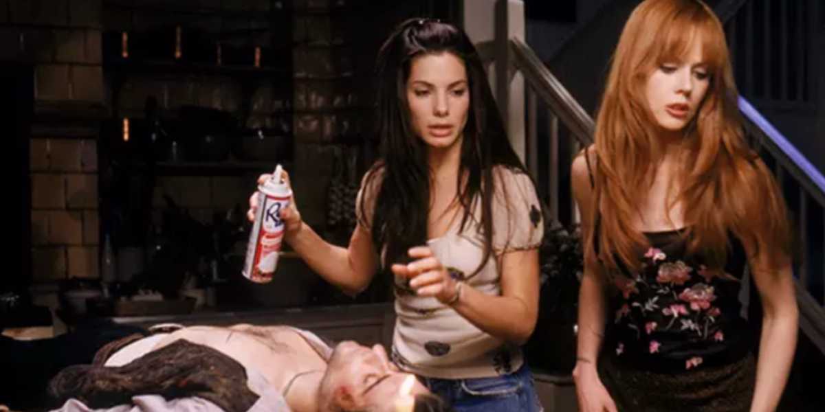 Two women are in a dimly lit room standing over an unconscious person lying on a table. One woman with dark hair is holding a spray can, and the other with light red hair looks surprised. The background suggests it is a kitchen or dining area.