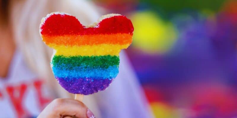 A person holds a Mickey Mouse-shaped treat covered in colorful, rainbow sugar. The stripes of color go from red, orange, yellow, green, blue, to purple. The background is blurred, with various bright colors and hints of a festive environment.
