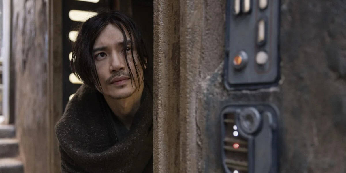 An individual with long dark hair and a mustache peeks around a corner in a weathered, industrial-looking setting. The person appears cautious or curious, looking towards a nearby control panel on the wall. The scene has a gritty, dystopian feel.