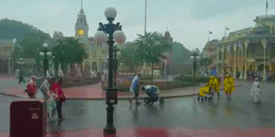 Guests are walking in Disney World on a rainy day. Some hold umbrellas or wear rain ponchos. A clock tower and colorful buildings are in the background. Trees and street lamps add to the scene. One person is pushing a stroller while others walk nearby, enjoying their trips despite the weather.
