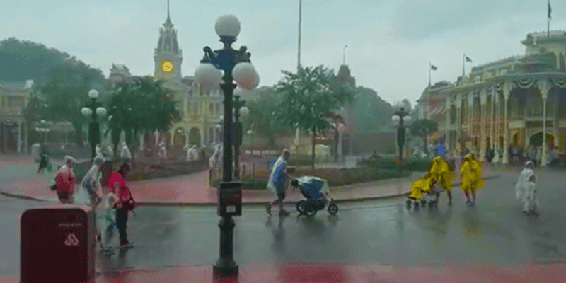 Guests are walking in Disney World on a rainy day. Some hold umbrellas or wear rain ponchos. A clock tower and colorful buildings are in the background. Trees and street lamps add to the scene. One person is pushing a stroller while others walk nearby, enjoying their trips despite the weather.