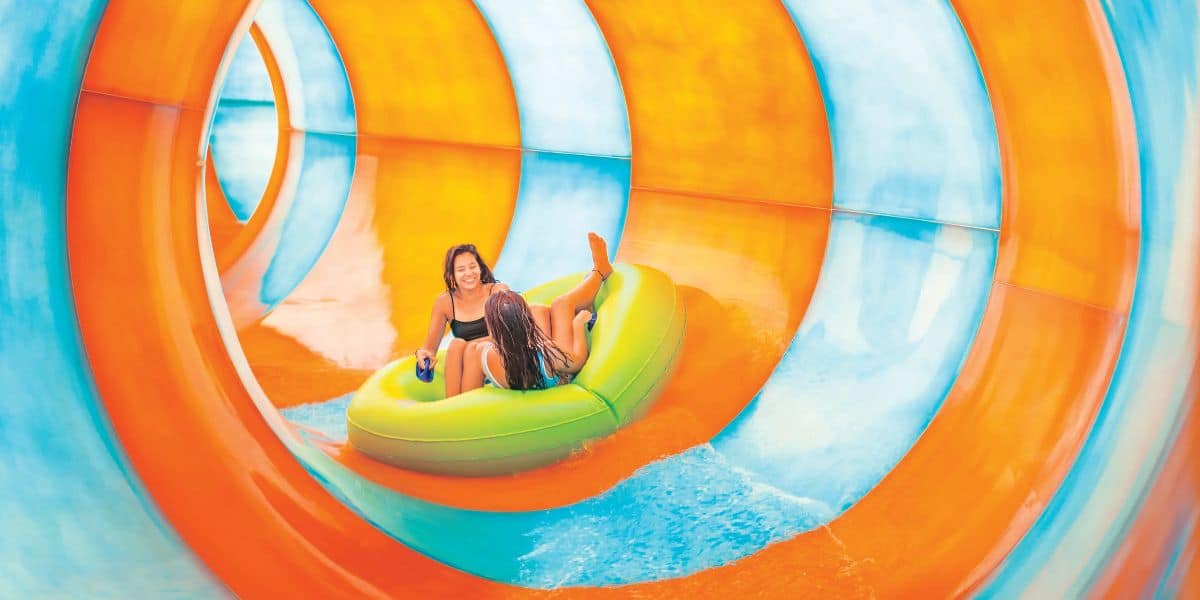 Two people ride a green inflatable raft through a brightly colored water slide at Florida water park Aquatica. The slide features orange, blue, and white sections that spiral as they glide down. Both riders appear to be enjoying the ride, surrounded by splashing water.