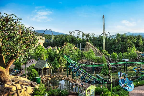 A vibrant amusement park with various roller coasters and rides set against a backdrop of forested hills and a clear blue sky. Thrill seekers are riding a green roller coaster, and numerous other attractions are visible, including a drop tower and ferris wheel.