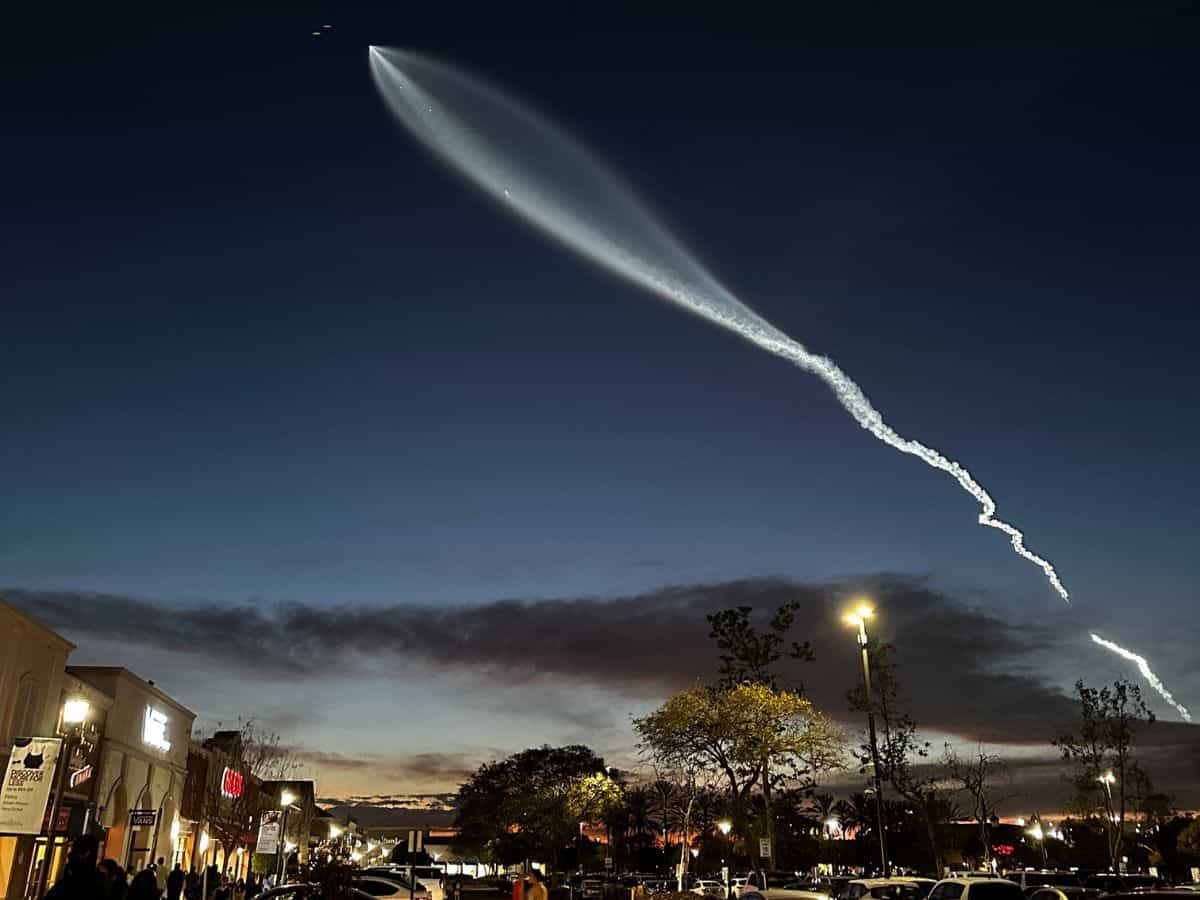 A long, illuminated trail arches across the evening sky, caused by the launch of Elon Musk's SpaceX rocket. The glow contrasts against the deep blue sky, with people gathered on the ground and images of buildings and streetlights visible below.