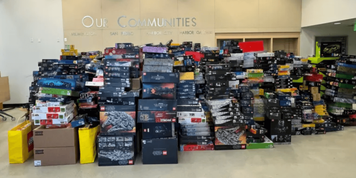 A large collection of boxed LEGO sets is stacked high against a wall in a room. The boxes are of various sizes and depict different themes and models. The wall has the words "Our Communities" and the names of various places written on it, almost as if stolen from the imagination of many diverse creators.