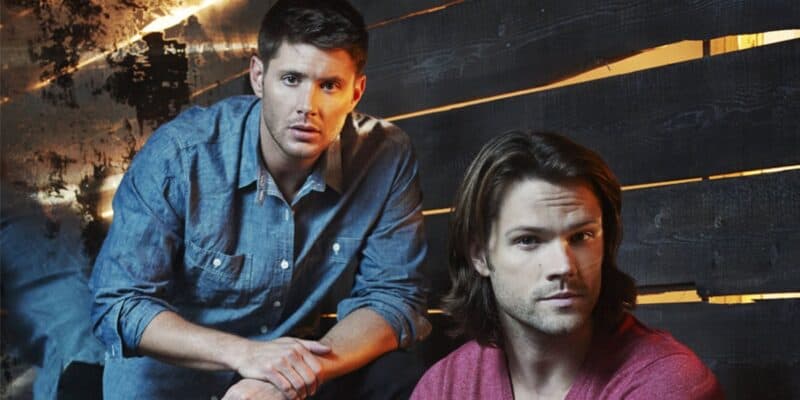 Jensen Ackles and Jared Padalecki as their Supernatural characters are shown indoors against a dark wooden backdrop. One man with short hair in a blue denim shirt is kneeling on one knee, while the other with long hair in a red shirt sits on a step. Both look directly at the camera with serious expressions.