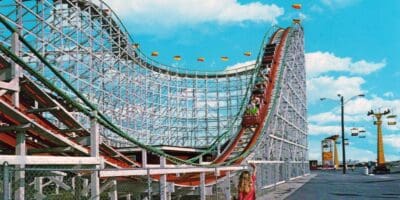 A vibrant amusement park roller coaster, featuring a large wooden structure with a steep drop, promises thrills but also carries risks. A red coaster car filled with people ascends the track. Bright blue sky and scattered clouds create a cheerful backdrop. Fenced pathway and cable cars visible in the background.