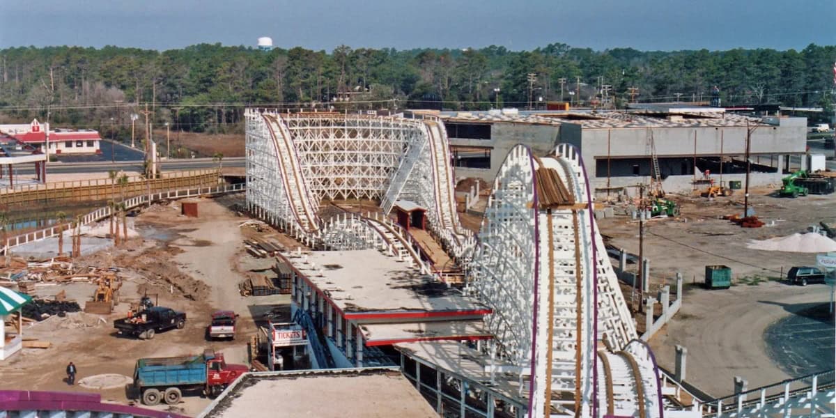 A construction site featuring a partially built wooden roller coaster with white supports and a red track. Various construction vehicles and materials surround the area, hinting at the precision needed to avoid creating a dangerous roller coaster. Trees and a few buildings are visible in the background.