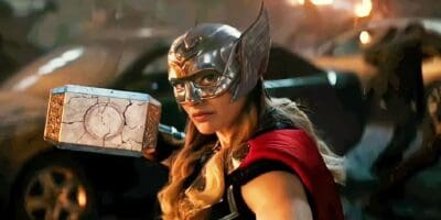 Natalie Portman in an elaborate superhero costume, including a helmet with wings and a red cape, holds a large, detailed hammer with both hands. The background appears to be a chaotic scene with dark, fiery elements.