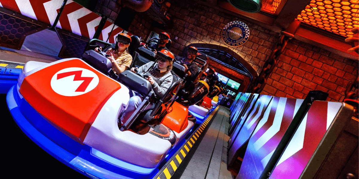 A group of people riding a Mario Kart-themed amusement park ride at Universal Studios, wearing virtual reality headsets and holding steering wheel controllers. The ride features colorful kart vehicles with the iconic Mario logo and a vibrant, animated environment.