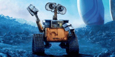 A rusty, yellow robot with large binocular-like eyes, named WALL-E, waves with one of its mechanical arms. The robot stands on rugged terrain with scattered debris, with a backdrop of a cloudy blue sky and large futuristic structures.