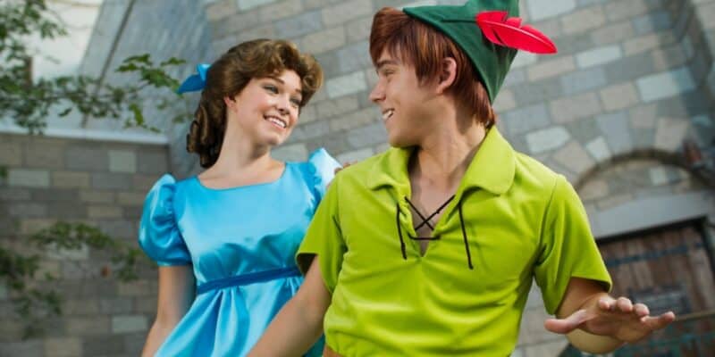 Two people dressed as Disney characters, Wendy and Peter Pan, smile at each other in an outdoor setting resembling a castle courtyard. Wendy wears a blue dress, while Peter Pan wears his iconic green outfit and hat with a red feather.