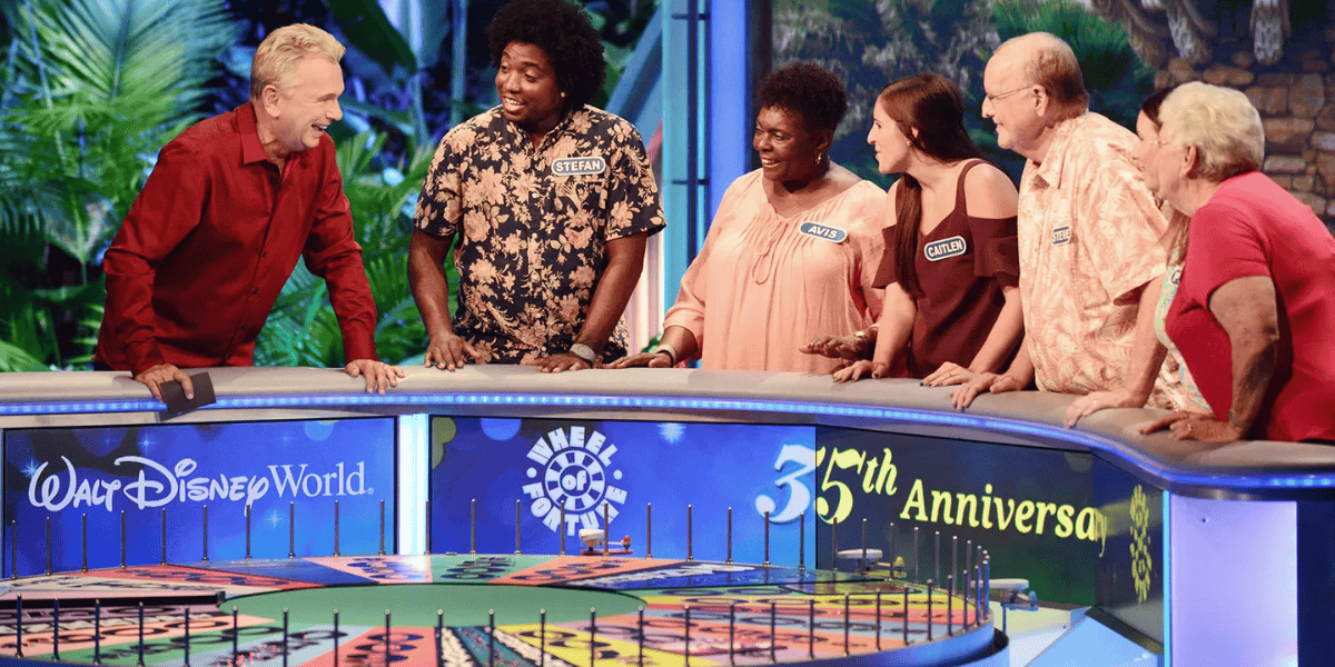 Six contestants are gathered around the Wheel of Fortune game show set at Walt Disney World during the show's 35th anniversary. They are smiling and engaged in conversation, celebrating a wild 43-year tradition. The colorful wheel is in the foreground, and the set has a festive atmosphere.