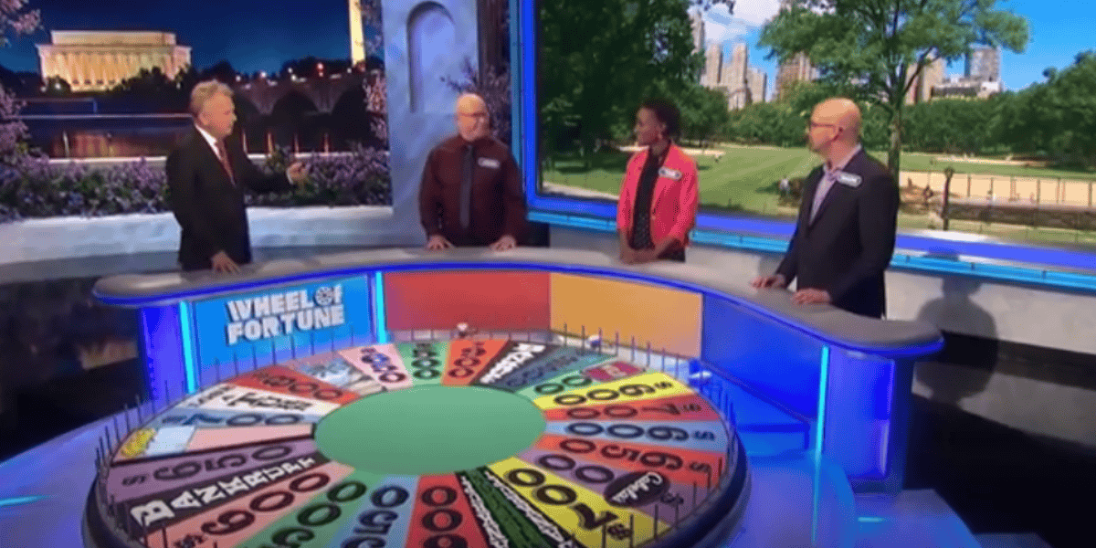 Three contestants stand behind podiums on the set of the game show "Wheel of Fortune," a wild 43-year tradition cherished by millions of fans. The colorful wheel is in the foreground with various numbers and slots, while the host stands on the left, engaging with the contestants. The backdrop shows an outdoor scene with trees and buildings.